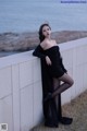 A woman in a black dress leaning against a wall by the ocean.